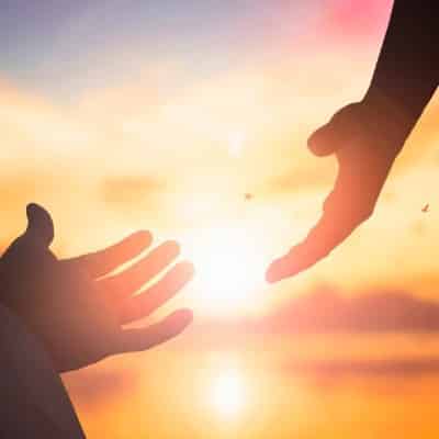 Zwei Hände, die einander reichen wollen, im Hintergrund die Sonne am Horizont.